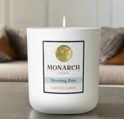 Monarch Candle labeled “Morning Rain” glowing gently in a white luxury glass vessel against a warm indoor background.