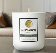 Monarch Candle labeled “Whispering Pines” glowing gently in a white luxury glass vessel against a warm indoor background.