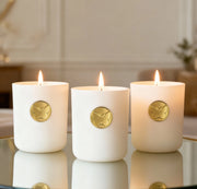 Three Monarch Candle votives glowing softly in a white luxury glass vessel with a gold butterfly charm on the front, placed on a reflective table in a warm indoor setting.
