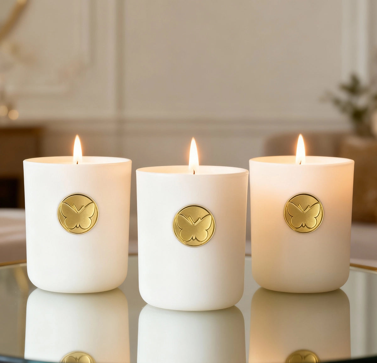 Three Monarch Candle votives glowing softly in a white luxury glass vessel with a gold butterfly charm on the front, placed on a reflective table in a warm indoor setting.
