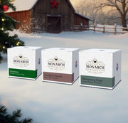 Three Monarch Candle votive boxes in front of a snowy barn with Christmas tree decorations. The boxes are labeled by scent: "Mistletoe", "Northern Hearth", and "Whispering Pines."