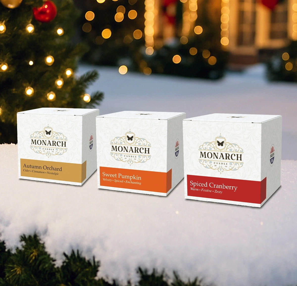 Three Monarch Candle Company votive boxes in front of a Christmas tree with snow. The boxes are labeled by scent: "Autumn Orchard", "Sweet Pumpkin", and "Spiced Cranberry"